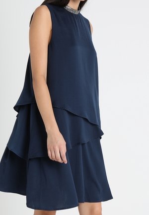 Woman wearing sleeveless navy blue layered dress with embellished high neckline against plain white background.