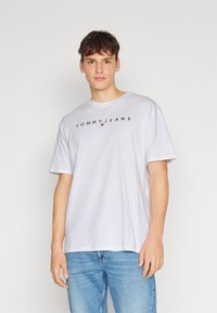 Baltas trumpa rankovė marškinėliai, pagaminti iš medvilnės, su juodu "TOMMY JEANS" logotipu per krūtinę. Derinami su mėlynomis džinsinėmis kelnėmis.