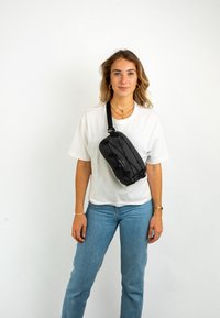 Czarna torba na ramię o prostokątnym kształcie, z teksturowaną powierzchnią, regulowanym paskiem i detalem z logo, noszona krzyżowo na białej koszuli.