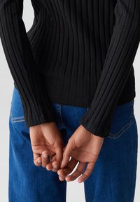 Schwarzes, geripptes Langarmoberteil, kombiniert mit blauen Denim-Jeans. Das Oberteil zeichnet sich durch ein figurbetontes Design mit strukturierter Stoffqualität und einem runden Halsausschnitt aus.