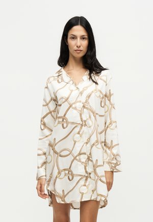 Vrouw met lang donker haar, die een witte overhemdjurk draagt met een beige riem en ringpatroon, staande tegen een effen lichte achtergrond.