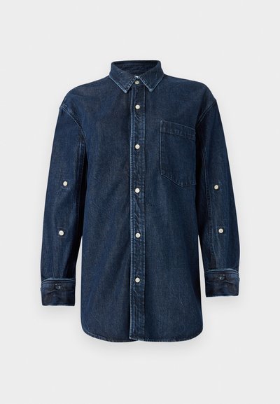 Chemise en denim bleu foncé avec un col boutonné, des manches longues, une poche avant et des boutons blancs. Le tissu a une apparence texturée.