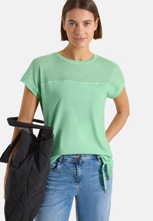 Femme portant un haut vert menthe à manches courtes et un jean bleu, tenant un sac matelassé noir sur son épaule gauche.