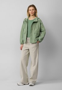 Lichtgroene gewatteerde jas met capuchon, elastische manchetten, ritssluiting en twee voorzakken, gecombineerd met lichtbeige rechte broek en grijze sneakers.