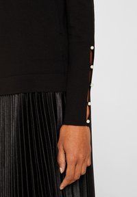 Černý pletený top s dlouhými rukávy a perlovými knoflíky podél bočních rozparek. Texturovaná černá plisovaná sukně částečně viditelná dole.