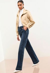 Lichtbeige cropped blazer met knoopsluiting, gecombineerd met donkerblauwe rechte jeans. Model draagt een witte coltrui en witte hakken.