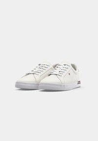 Baskets basses blanches avec lacets blancs, petit logo sur le côté, et un détail rayé sur la semelle, présentées sur un fond uni.