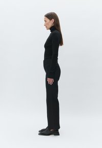 Black turtleneck sweater and fitted black trousers paired with black shoes; model stands in profile against a plain white background.