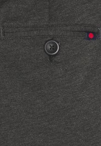 Donkergrijze stof met een voorklep met een zwarte knop en een accentlabel in rood en marineblauw. Geweven materiaal met een subtiele afwerking.