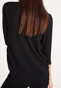 Blusa nera trasparente con maniche a tre quarti e vestibilità ampia. Tessuto liscio; la vista dal retro mostra i capelli che scendono oltre le spalle.