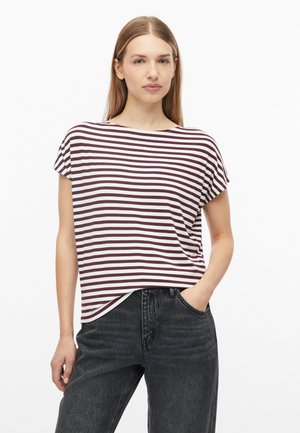 Jonge vrouw met steil haar die een wit met donkerrood horizontaal gestreept topje met korte mouwen en een donker, hooggesloten spijkerbroek draagt.