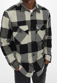 Czarna i biała koszula flanelowa w kratę z kołnierzykiem zapinanym na guziki, dwoma kieszeniami na piersi i fakturowanym materiałem. Szare dżinsy dopełniają stylizację.