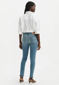 Vaqueros ajustados de color azul en un lavado medio con cintura alta, bolsillos traseros con detalles de costura, combinados con una camisa blanca de botones.