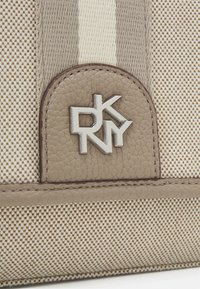 Beige handbag with textured fabric and smooth leather accents. Features a silver DKNY logo, striped design, and a woven pattern.