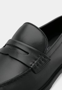 Mocassins en cuir noir à finition lisse, avec une couture décorative et une bride centrale surélevée à l'avant. Semelle plate.