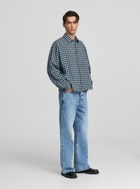 Camicia a quadri con bottoni nei colori blu, bianco e marrone; taglio comodo. Jeans in denim azzurro chiaro, taglio largo, abbinati a calzature nere.