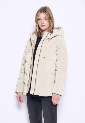 Giacca imbottita beige con cappuccio, chiusura con zip e polsini elasticizzati; texture trapuntata con cuciture diagonali; indossata sopra una maglietta nera.