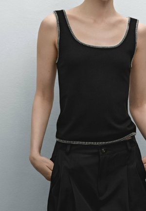 Person iført en ærmeløs sort top med hvide sting og sorte plisserede bukser med hænderne i lommerne, neutralt baggrund.