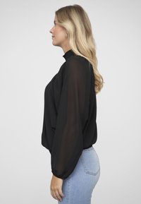 Schwarze transparente Bluse mit langen, puffigen Ärmeln und einem gerüschten, hohen Kragen, kombiniert mit hellblauen, eng anliegenden Jeans. Glatte Textur, fließendes Design.