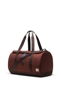 Sac de voyage brun de forme cylindrique. Fabriqué en tissu robuste, il possède des bretelles marron foncé, une tirette bleue et un écusson avec logo.