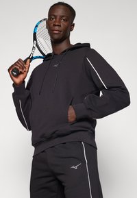 Black hoodie with white side stripes, front pocket, and drawstring. Worn with matching black pants, holding a tennis racket.