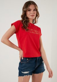 Rød bomulds t-shirt med korte ærmer og rund halsudskæring, prydet med guldtekst, der læser "no love lost" i et gentaget mønster. Parret med denimshorts.