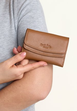 Brown leather wallet with a textured finish, featuring a curved flap and embossed branding, held against a gray fabric background.