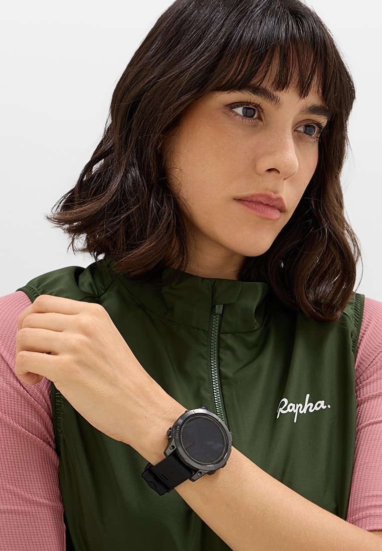 Reloj inteligente negro en una muñeca, cara redonda, acentos de metal resistente, correa texturizada negra. Chaleco verde con cremallera y logo blanco, detalle de manga rosa.