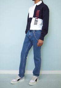 Polo de manga larga en azul marino y blanco con texto rojo, combinado con unos vaqueros azul claro y zapatillas beiges. Presenta un detalle de cadena en los vaqueros.