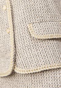 Biała i czarna marynarka z tkaniny tweed, o fakturowanym materiale, z akcentami z złotej nici i okrągłymi, złotymi guzikami. Posiada plecioną lamówkę oraz kieszeń z klapą.