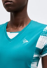 Κοντινό πλάνο ατόμου που φοράει αθλητική μπλούζα από πετρόλ με λαιμόκοψη V και λευκά γεωμετρικά σχέδια στα μανίκια, καθώς και μικρό λογότυπο στο στήθος.