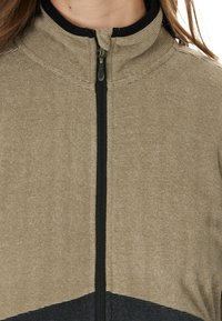 Veste zippée en beige avec des bandes horizontales texturées et un accent noir en bas. Elle dispose d'un col montant et d'une fermeture éclair à l'avant.