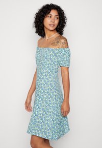 Vestido floral, design à tona dos ombros, padrão azul claro e verde. Mangas bufantes curtas, corpete justo, saia fluida com uma bainha levemente rodada.