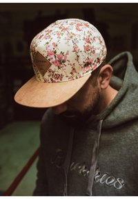Cappello floreale con sfondo crema e fiori rosa, dotato di visiera in suede e un patch a forma di diamante. Felpa grigia scura visibile.
