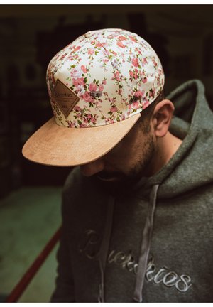 Gorra floral con fondo crema y flores rosas, con visera de ante y un parche de logo en forma de diamante. Sudadera gris oscuro visible.