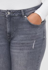 Jeans de ganga cinzentos com uma cintura de altura média, cinco bolsos e um detalhe desgastado perto da coxa. Textura suave com desvanecimento subtil.