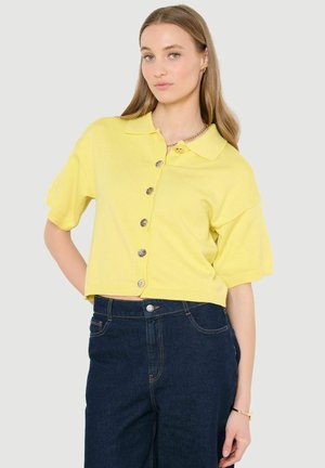 Mladá žena v žltom krátkom nátelníku s gombíkmi a tmavomodrých džínsach s vysokým pásom pózuje s jednou rukou na bokoch.