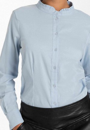Femme portant une chemise bleu clair à manches longues boutonnée avec un col volanté et un pantalon en cuir noir, mains dans les poches.