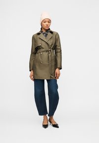 Γυναίκα με ανοιχτό ροζ σκουφάκι, χακί παλτό με ζώνη, καρό πουκάμισο, φαρδιά σκουρό μπλε τζιν και μαύρα μυτερά τακούνια, που στέκεται σε λευκό φόντο.