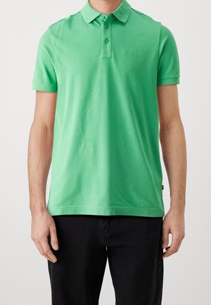 Homme portant un polo vert vif à manches courtes avec un pantalon noir sur un fond clair uni.