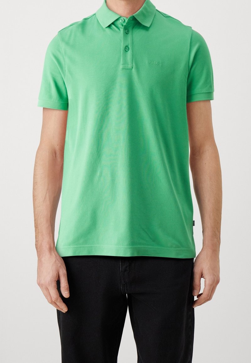 Homme portant un polo vert vif à manches courtes avec un pantalon noir sur un fond clair uni.