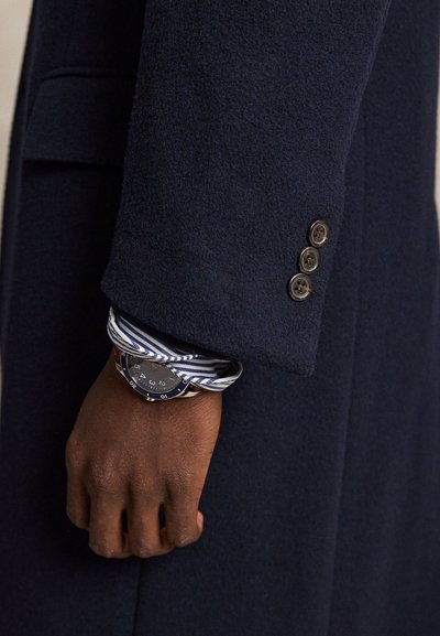 Main à la peau foncée portant une chemise rayée et une montre, sortant d'une manche de manteau bleu marine avec trois boutons au poignet.
