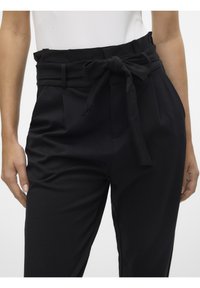 Pantalons noirs taille haute avec une ceinture en tissu, présentant des plis et une texture lisse. Vue en gros plan, mains tenant la taille.