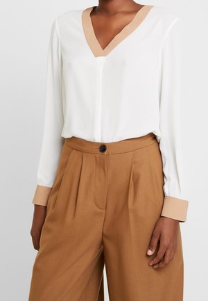 Femme portant un chemisier blanc à col et poignets beige rentré dans un pantalon large taille haute couleur camel avec un bouton noir.