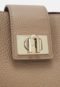 Patte de sac en cuir beige texturé avec un fermoir rectangulaire doré comportant un détail métallique courbé et le nom de la marque Furla.