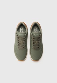 Adidași verzi cu un superior sintetic neted, accente perforate, design cu șireturi și o talpă din cauciuc maro. Numele brandului este vizibil pe limbă.
