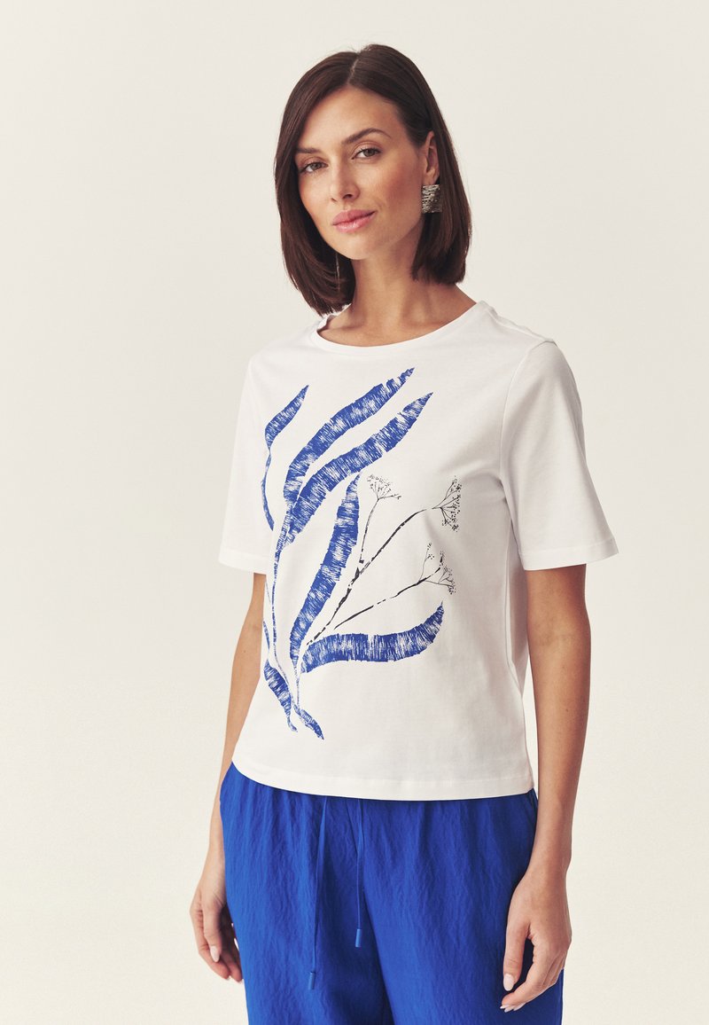 Femme portant une chemise blanche à manches courtes avec un imprimé abstrait de feuilles bleues et un pantalon bleu roi, debout devant un fond clair uni.