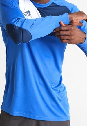 Mann passt den Ärmel eines blauen Adidas-Sportshirts mit dunklen Ellbogenschonern an, vor einem einfarbig weißen Hintergrund.
