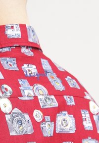Red short-sleeve shirt featuring a camera print in various colors. The fabric is lightweight with a smooth texture.