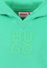 Groene hoodie met ribbelstructuur, voorzien van een groot reliëf "HUGO" logo in het midden. Details van de capuchon en hals zichtbaar.